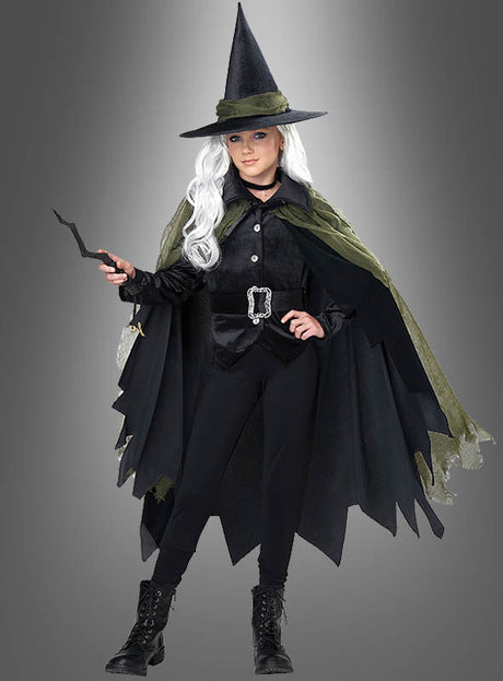 Ein Mädchen, das das Hexenkostüm „Layla für Mädchen“ in Schwarz und Grün trägt, dazu einen spitzen Hut, eine weiße Perücke und schwarze Stiefel, und einen schwarzen Zauberstab in der Hand hält, steht vor einem grauen Hintergrund.