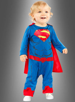 Ein Kleinkind, das das Superman-Kostüm für Kleinkinder trägt – einen blauen Anzug mit rotem Umhang –, steht vor einem grauen Hintergrund und lächelt, während es stolz sein Original-Lizenzkostüm präsentiert.