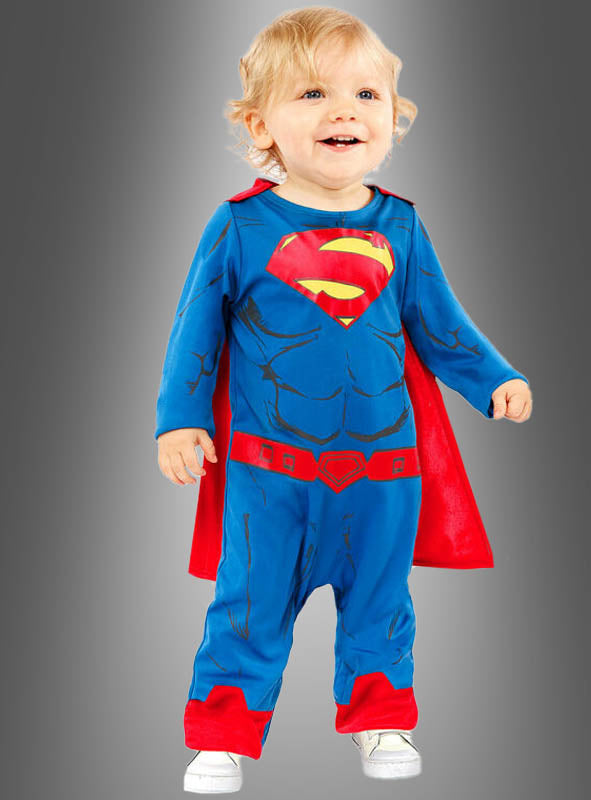 Ein Kleinkind, das das Superman-Kostüm für Kleinkinder trägt – einen blauen Anzug mit rotem Umhang –, steht vor einem grauen Hintergrund und lächelt, während es stolz sein Original-Lizenzkostüm präsentiert.