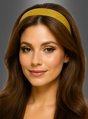 Eine Frau mit langen braunen Haaren trägt das goldene Glitzer-Haarband „Disco Glitzer“, dazu ein dezentes Make-up und ein leichtes Lächeln, vor einem grauen Hintergrund.