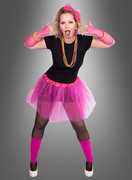 Eine Person, die ein 80er-Jahre-Kostüm-Set mit Tüllrock trägt – bestehend aus einem schwarzen Shirt, einem rosa Tutu, rosa Handschuhen, Perlenketten, rosa Stulpen und weißen Pumps –, steht vor einem grauen Hintergrund und schneidet eine verspielte Grimasse.