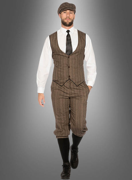 Ein Mann, der das „20er-Jahre-Mode“-Kostüm „John“ trägt – bestehend aus einer im Vintage-Stil gehaltenen braunen Weste, einer dazu passenden Knickerbocker-Hose, einem weißen Hemd, einer schwarzen Krawatte, einer Schirmmütze, schwarzen Socken und schwarzen Schuhen –, verkörpert den unverwechselbaren „Peaky Blinders“-Look, während er auf die Kamera zugeht.