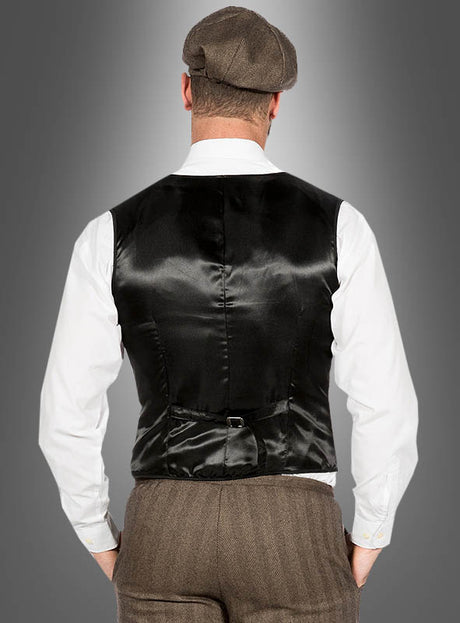 Ein Mann, der das „20er-Jahre-Modekostüm John“ trägt – bestehend aus einer schwarzen Satinweste, einem weißen Hemd, einer braunen Hose und einer Schirmmütze –, steht mit dem Rücken zur Kamera vor einem grauen Hintergrund mit Farbverlauf und verkörpert damit den klassischen „Peaky Blinders“-Stil sowie die zeitlose Mode der 1920er Jahre.