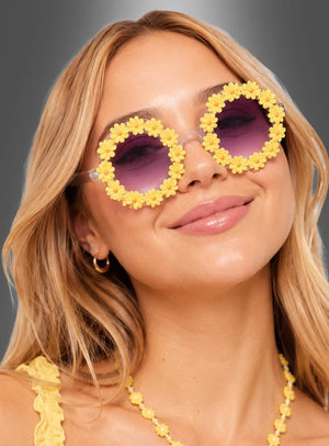 Eine Frau mit langen blonden Haaren trägt eine runde „Hippie Blumen“-Sonnenbrille mit gelbem Blumenrahmen und violetten Gläsern, dazu eine gelbe Halskette; sie posiert vor einem grauen Hintergrund.