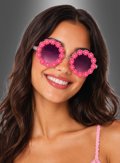 Eine Frau mit langen braunen Haaren lächelt, während sie die „Hippie Blumen“-Sonnenbrille – eine große, runde Sonnenbrille, die mit rosa Blumen verziert ist – und ein rosa Oberteil trägt; im Hintergrund ist ein grauer Farbverlauf zu sehen.