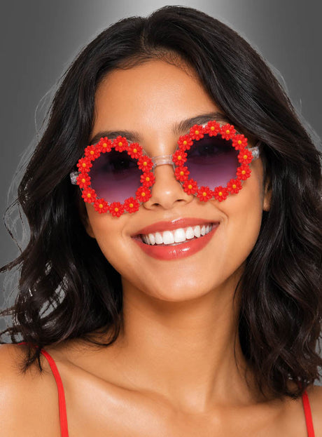 Eine lächelnde Frau trägt die „Hippie Blumen“-Sonnenbrille – eine runde Sonnenbrille, die mit roten und gelben Blumenmotiven verziert ist und an den klassischen Flower-Power-Stil erinnert. Ihr dunkles, welliges Haar umrahmt ihr Gesicht, und auf ihrer Schulter ist ein roter Riemen zu sehen, der sich vor einem grauen Hintergrund abhebt.