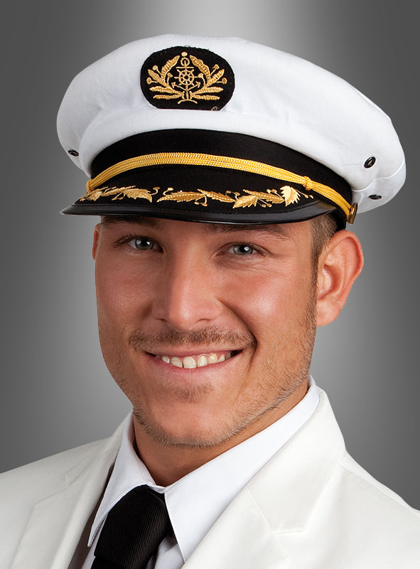 Ein Mann, der eine Kapitänsmütze für Marineoffiziere trägt – weiß mit einem markanten schwarz-goldenen Emblem –, dazu eine weiße Jacke und eine schwarze Krawatte, lächelt selbstbewusst vor einem grauen Hintergrund mit Farbverlauf und verkörpert die souveräne Ausstrahlung eines erfahrenen Marineoffiziers.
