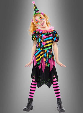 Ein Kind, das das „Zirkus Clown Neon Kostüm Mädchen“ trägt – mit gestreiften Strumpfhosen, einem farbenfrohen, gemusterten Kleid, einem Partyhut, Gesichtsbemalung und Stiefeln – steht vor einem grauen Hintergrund und eignet sich somit perfekt für Kinderfasching-Feiern.