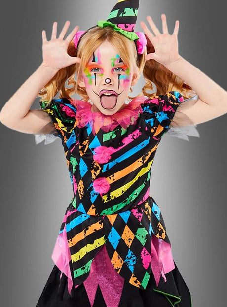 Ein Kind, das das farbenfrohe „Zirkus Clown Neon Kostüm Mädchen“ und den dazu passenden Hut trägt, macht eine verspielte Grimasse: Die Hände sind erhoben, die Zunge herausgestreckt und das Gesicht ist bunt bemalt – vor einem grauen Hintergrund mit Farbverlauf – perfekt für die Kinderfasching-Feierlichkeiten.