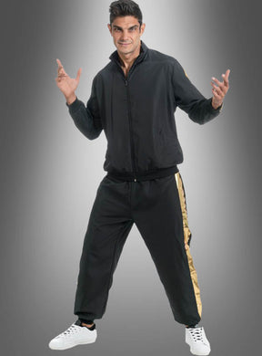 Ein Mann steht lächelnd vor einem grauen Hintergrund mit Farbverlauf und trägt den Rapper-Trainingsanzug in Schwarz-Gold – eine schwarze Jacke mit Reißverschluss und eine dazu passende Hose mit goldenen Streifen – sowie weiße Turnschuhe.