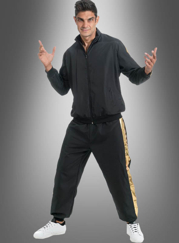 Ein Mann steht lächelnd vor einem grauen Hintergrund mit Farbverlauf und trägt den Rapper-Trainingsanzug in Schwarz-Gold – eine schwarze Jacke mit Reißverschluss und eine dazu passende Hose mit goldenen Streifen – sowie weiße Turnschuhe.