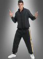 Ein Mann steht lächelnd vor einem grauen Hintergrund mit Farbverlauf und trägt den Rapper-Trainingsanzug in Schwarz-Gold – eine schwarze Jacke mit Reißverschluss und eine dazu passende Hose mit goldenen Streifen – sowie weiße Turnschuhe.