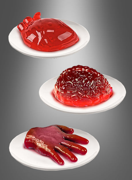 Auf drei weißen Tellern liegen Gelatine-Desserts in Form eines Herzens, eines Gehirns und einer Hand – alle in leuchtendem Rot vor einem grauen Hintergrund mit Farbverlauf. Diese gruseligen Leckereien wurden mit den Halloween-Puddingformen „Herz“, „Gehirn“ oder „Hand“ hergestellt und umfassen einen wackeligen „Gehirn-Wackelpudding“ sowie ein auffälliges „Gelatine-Herz“ – perfekt für eine unheimliche Halloween-Dekoration.