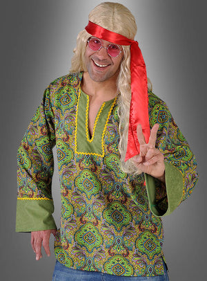 Ein Mann, der ein „Grünes Hippiehemd“ im „Summer of Love“-Stil als 70er-Jahre-Kostüm trägt, dazu eine lange blonde Perücke, ein rotes Stirnband und eine rosa Brille, lächelt und formt mit der rechten Hand das Peace-Zeichen.