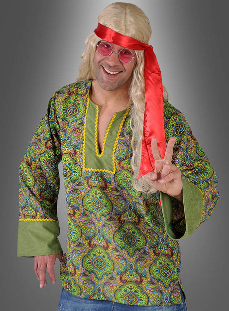 Ein Mann, der ein „Grünes Hippiehemd“ im „Summer of Love“-Stil als 70er-Jahre-Kostüm trägt, dazu eine lange blonde Perücke, ein rotes Stirnband und eine rosa Brille, lächelt und formt mit der rechten Hand das Peace-Zeichen.