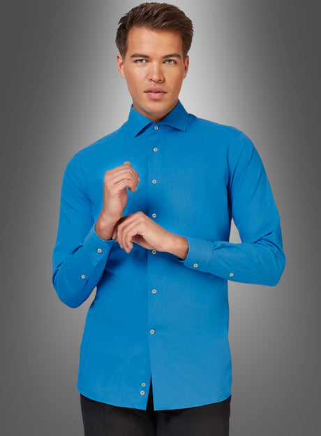 Ein Mann, der ein blaues Hemd im klassischen Stil und eine schwarze Hose trägt, steht vor einem grau verlaufenden Hintergrund und blickt mit den Händen vor der Brust nach vorne.