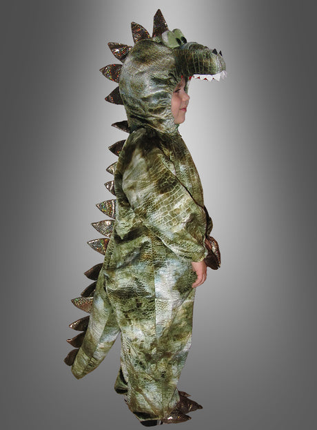 Ein Kind, das das Dinosaurier-Kostüm für Kinder trägt – aus grünem Stoff mit Stacheln am Rücken –, steht im Profil vor einem grau verlaufenden Hintergrund.