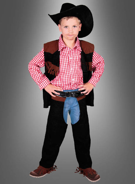 Ein kleiner Junge im Cowboy-John-Kinderkostüm – mit schwarzem Hut, rotem Karohemd, schwarzer Weste, Chaps und braunen Schuhen – steht mit den Händen in die Hüften gestemmt vor einem grauen Hintergrund mit Farbverlauf.