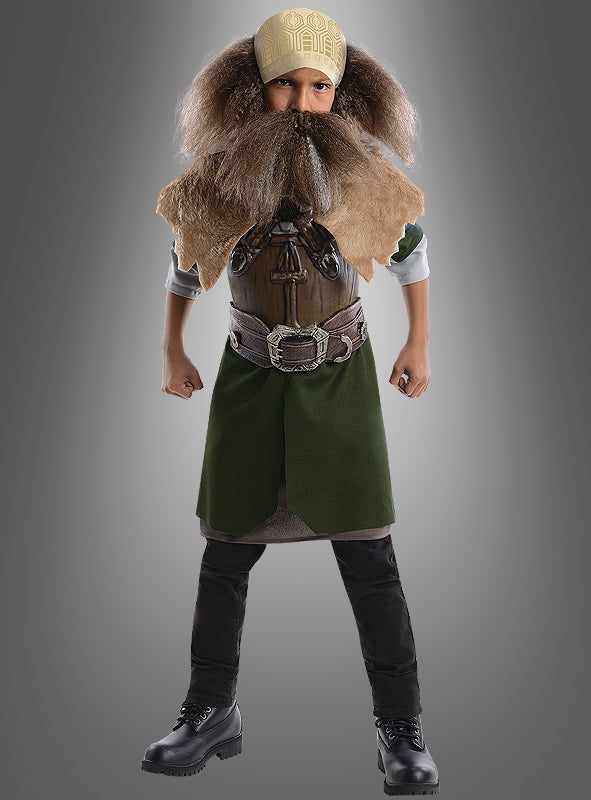 Ein Kind, das das Dwalin-Kinderkostüm aus „Der Hobbit“ trägt – darunter einen großen Kunstbart, eine braune Weste, eine grüne Tunika, schwarze Stiefel und ein gelb gemustertes Stirnband –, steht mit angewinkelten Armen und geballten Fäusten da.
