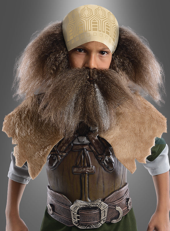 Ein Kind, das das Dwalin-Kinderkostüm aus „Der Hobbit“ trägt – bestehend aus einem großen Kunstbart, einem Pelzumhang und einem gemusterten Kopftuch –, posiert vor einem grauen Hintergrund.