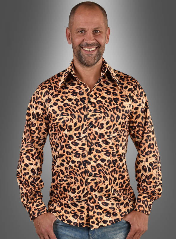 Ein Mann mit Glatze lächelt, während er das „Herrenhemd Leoprint 80er Jahre“ trägt – ein langärmeliges Hemd mit Knopfleiste und auffälligem Leopardenmuster –, dazu blaue Jeans, vor einem grauen Hintergrund mit Farbverlauf.