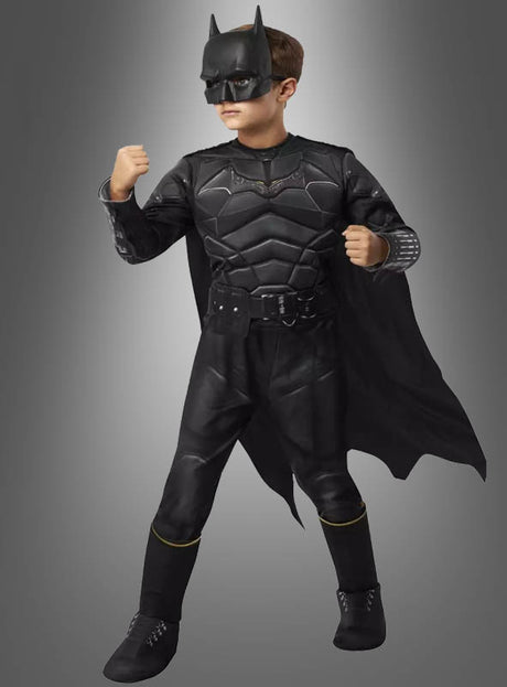 Ein Kind, das ein „Batman Luxus“-Kinderkostüm trägt – komplett mit Umhang, Maske und Handschuhen –, steht mit erhobenen Fäusten vor einem schlichten Hintergrund mit Farbverlauf.