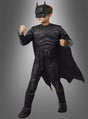 Ein Kind, das ein „Batman Luxus“-Kinderkostüm trägt – komplett mit Umhang, Maske und Handschuhen –, steht mit erhobenen Fäusten vor einem schlichten Hintergrund mit Farbverlauf.