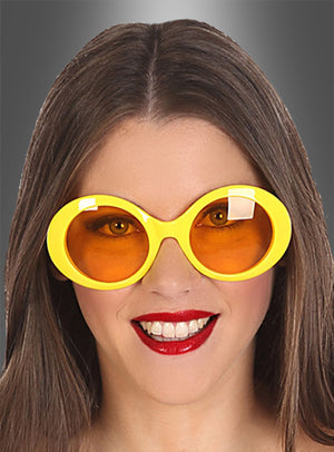 Eine Frau mit langen braunen Haaren lächelt. Sie trägt die „Brille im 60er-Jahre-Retro-Look“ – eine große, runde gelbe Sonnenbrille mit orangefarbenen Gläsern – sowie roten Lippenstift und posiert vor einem grauen Hintergrund mit Farbverlauf.