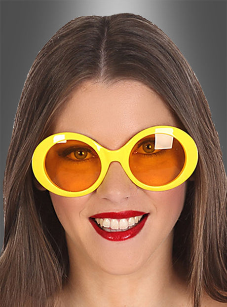 Eine Frau mit langen braunen Haaren lächelt. Sie trägt die „Brille im 60er-Jahre-Retro-Look“ – eine große, runde gelbe Sonnenbrille mit orangefarbenen Gläsern – sowie roten Lippenstift und posiert vor einem grauen Hintergrund mit Farbverlauf.