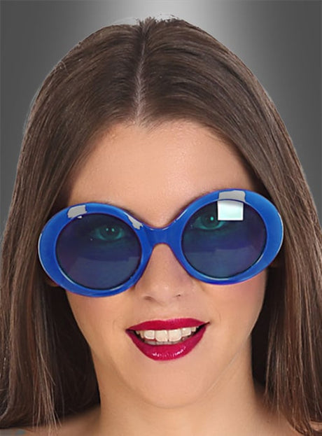 Eine Person mit glattem braunem Haar trägt eine große, runde blaue Brille im Retro-Look der 60er Jahre und roten Lippenstift und lächelt vor einem grauen Hintergrund mit Farbverlauf.