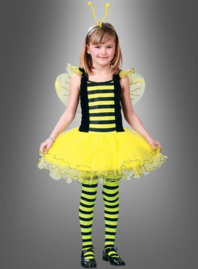 Ein kleines Mädchen steht mit dem Blick nach vorne und trägt das „Emsiges Bienchen“-Kinderkostüm mit gelben und schwarzen Streifen, zarten Flügeln, einem Tutu-Rock, gestreiften Leggings und einem Stirnband mit Fühlern vor einem schlichten grauen Hintergrund. Dieses bezaubernde „Kinderkostüm Biene“ eignet sich perfekt für Fasching oder zum Verkleiden.