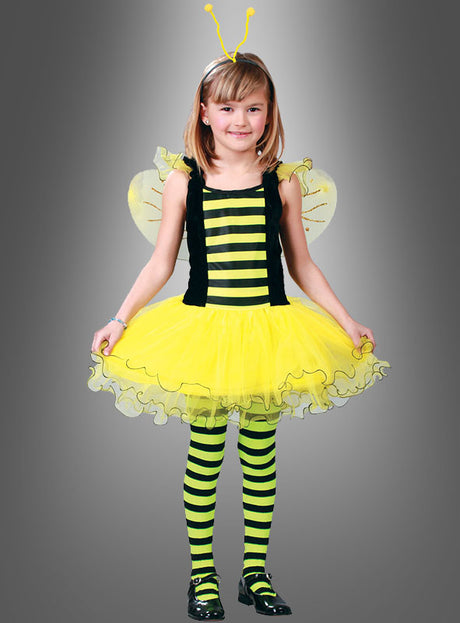 Ein kleines Mädchen steht mit dem Blick nach vorne und trägt das „Emsiges Bienchen“-Kinderkostüm mit gelben und schwarzen Streifen, zarten Flügeln, einem Tutu-Rock, gestreiften Leggings und einem Stirnband mit Fühlern vor einem schlichten grauen Hintergrund. Dieses bezaubernde „Kinderkostüm Biene“ eignet sich perfekt für Fasching oder zum Verkleiden.