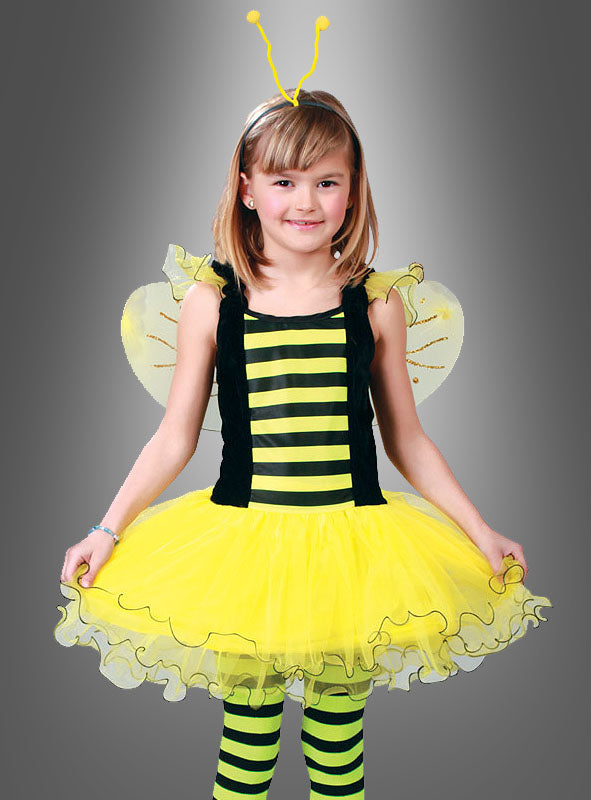 Ein kleines Mädchen trägt ein „Emsiges Bienchen“-Kinderkostüm – bestehend aus einem gelb-schwarz gestreiften Bienenkostüm, zierlichen Flügeln, einem gelben Tutu und einem Stirnband mit Fühlern – und steht vor einem grauen Hintergrund. Dieses bezaubernde Bienen-Kinderkostüm eignet sich ideal für Fasching oder jede andere Gelegenheit, bei der man sich verkleiden möchte.