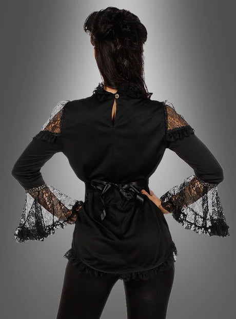 Eine Frau steht mit dem Rücken zur Kamera, die Hände in die Hüften gestemmt, und trägt die „Elegante Bluse mit Spitze, schwarz“ mit Rüschenverzierungen vor einem grauen Hintergrund mit Farbverlauf.