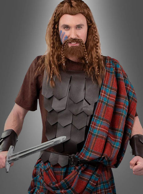 Ein Mann, der das „Highlander“-Schottenkostüm trägt – eine mittelalterliche Kriegerkleidung bestehend aus einem Brustpanzer, einer an einen Schottenrock angelehnten Tartan-Schärpe, geflochtenen Haaren, einem künstlichen Bart und Gesichtsbemalung, der ein Schwert in der Hand hält –, steht vor einem schlichten grauen Hintergrund.