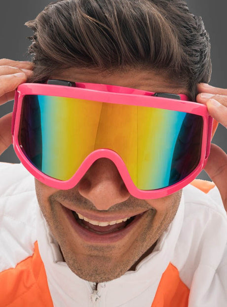 Eine Person in einer weiß-orangefarbenen Jacke lächelt, während sie eine große, pinkfarbene, reflektierende Skibrille „Karneval Neon“ zurechtzieht – ein auffälliges Après-Ski-Accessoire, das sich perfekt für festlichen Winterspaß eignet.