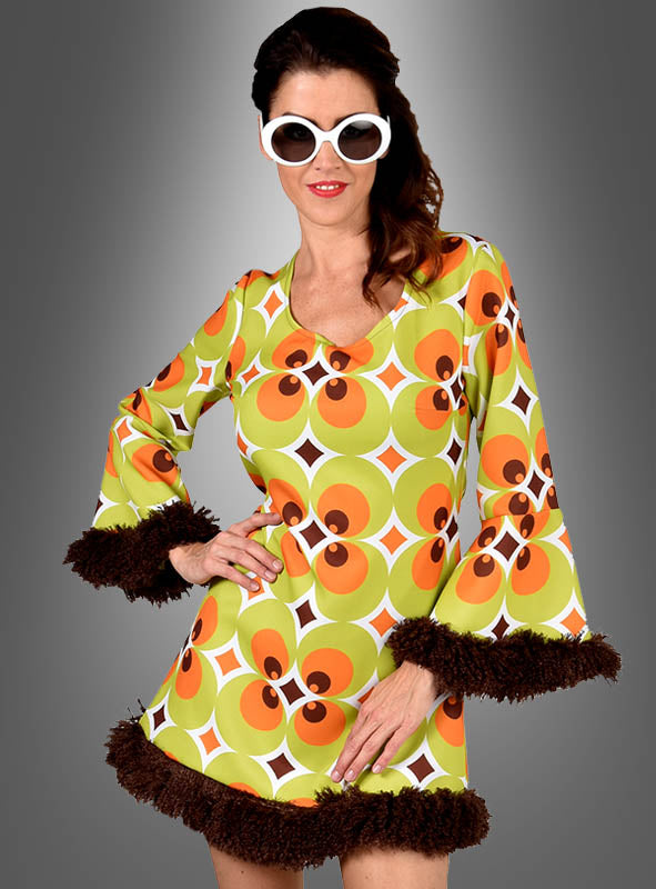 Eine Frau trägt das „70er Retrokleid Damen grün-braun“ – ein Retro-Kleid mit grünem, orangefarbenem und gelbem Muster sowie braunen Verzierungen –, dazu eine weiße Sonnenbrille, und posiert vor einem Hintergrund mit Farbverlauf. Dies ist die perfekte Wahl für jede Mottoparty oder eine Feier im Vintage-Stil.
