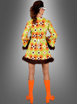 Eine Person in einem grün-braunen 70er-Jahre-Retro-Kleid für Damen mit grünen und braunen Retro-Mustern und braunen Fransen, orangefarbenen kniehohen Stiefeln und einer weißen Sonnenbrille – perfekt als 70er-Jahre-Damenkostüm – steht mit dem Rücken zur Kamera vor einem grauen Hintergrund mit Farbverlauf.