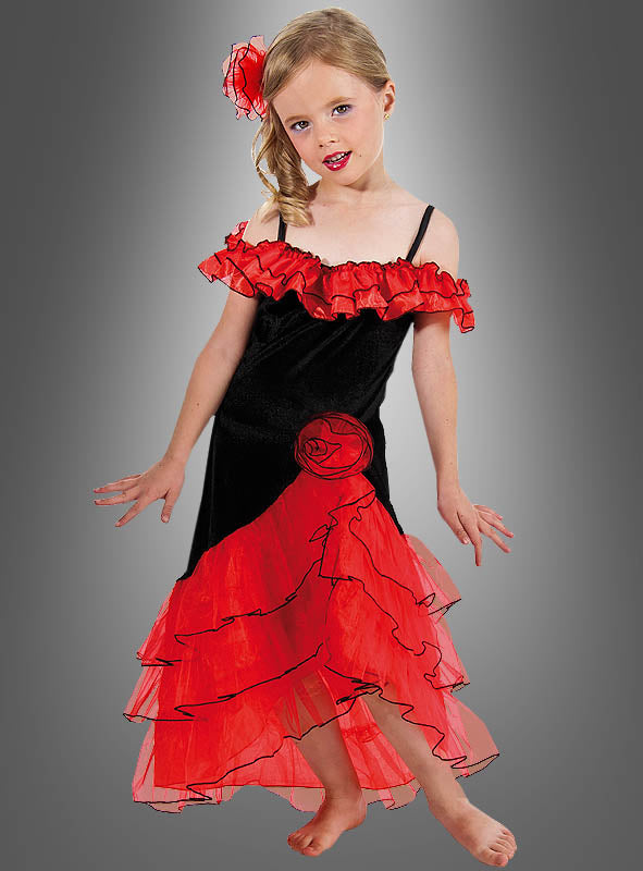 Ein junges Mädchen präsentiert das Spanierin-Kostüm für Kinder „Carmen“ – ein schwarz-rotes Rüschenkleid mit passendem Haarschmuck –, während sie barfuß vor einem grauen Hintergrund mit Farbverlauf steht.