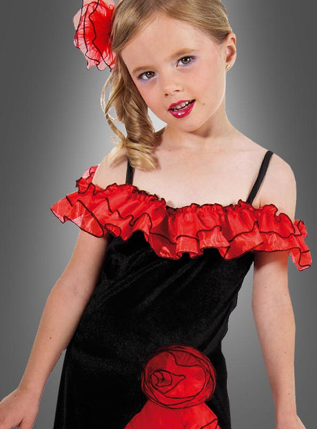 Ein junges Mädchen präsentiert das „Spanierin“-Kostüm für Kinder „Carmen“, bestehend aus einem schwarzen Kleid mit roten Rüschen und einem passenden roten Blumenaccessoire im Haar, vor einem grauen Hintergrund.