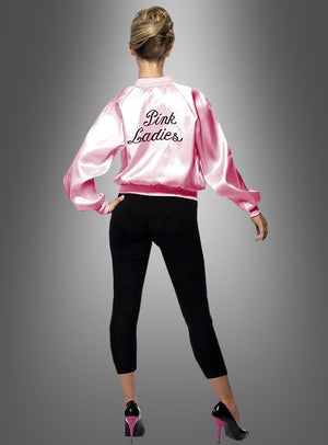 Eine Frau steht mit dem Rücken zur Kamera und trägt die „Pink Lady“-Jacke aus „Grease – 60er Jahre“, dazu schwarze Leggings und rosa High Heels, vor einem grauen Hintergrund mit Farbverlauf.