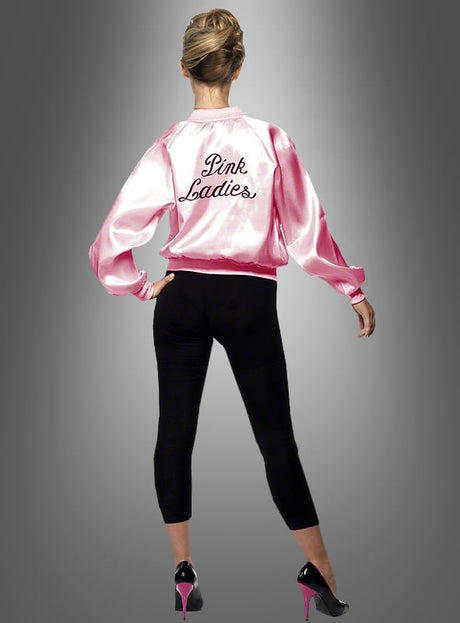 Eine Frau steht mit dem Rücken zur Kamera und trägt die „Pink Lady“-Jacke aus „Grease – 60er Jahre“, dazu schwarze Leggings und rosa High Heels, vor einem grauen Hintergrund mit Farbverlauf.
