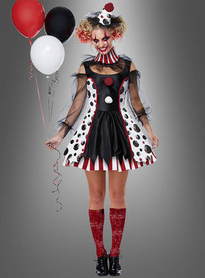 Eine Frau, die das Clown-Kleid „Pippa für Damen“ trägt, das sich durch lebhafte Polka-Dot- und Streifenmuster auszeichnet, hält rote, weiße und schwarze Luftballons in der Hand. Sie trägt auffälliges rotes Make-up und glitzernde rote Kniestrümpfe – ein ideales Karnevalskostüm für Halloween – und steht vor einem grauen Hintergrund.