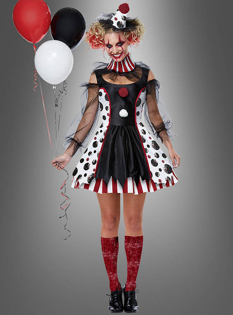 Eine Frau, die das Clown-Kleid „Pippa für Damen“ trägt, das sich durch lebhafte Polka-Dot- und Streifenmuster auszeichnet, hält rote, weiße und schwarze Luftballons in der Hand. Sie trägt auffälliges rotes Make-up und glitzernde rote Kniestrümpfe – ein ideales Karnevalskostüm für Halloween – und steht vor einem grauen Hintergrund.