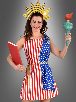 Eine Frau in einem von der Freiheitsstatue inspirierten „Stars & Stripes USA“-Flaggenkleid mit Sternen und Streifen, die ein rotes Buch und eine Fackel in der Hand hält und eine Krone mit Sonnenstrahlen trägt – dieses Kostüm eignet sich perfekt, um vor dem grauen Hintergrund ein patriotisches Statement zu setzen.