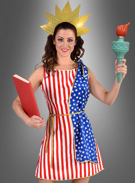 Eine Frau in einem von der Freiheitsstatue inspirierten „Stars & Stripes USA“-Flaggenkleid mit Sternen und Streifen, die ein rotes Buch und eine Fackel in der Hand hält und eine Krone mit Sonnenstrahlen trägt – dieses Kostüm eignet sich perfekt, um vor dem grauen Hintergrund ein patriotisches Statement zu setzen.
