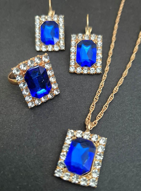 Ein raffiniertes Edles Schmuckset mit Edelsteinen und Strass, bestehend aus goldfarbenem Schmuck, der mit blauen, rechteckigen Edelsteinen verziert ist, die von funkelnden, klaren Steinen umrahmt werden – bestehend aus einem Ring, Ohrringen und einer Halskette –, vor schwarzem Hintergrund arrangiert. Diese elegante Kollektion strahlt raffinierten Stil aus und ist somit die ideale Wahl für jeden besonderen Anlass.