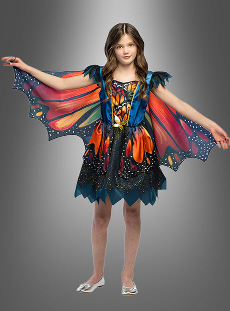Ein Mädchen steht mit ausgestreckten Armen da und trägt das „Kleid mit Schmetterlingsflügel“ mit flügelartigen Ärmeln und einem dazu passenden Rock, das vor einem grauen Hintergrund mit Farbverlauf wie ein wunderschöner Schmetterling wirkt.