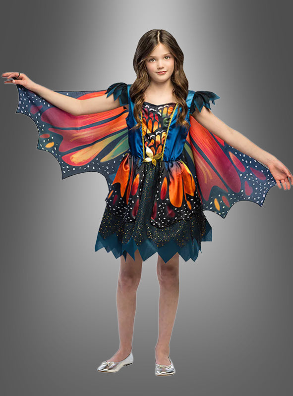 Ein Mädchen steht mit ausgestreckten Armen da und trägt das „Kleid mit Schmetterlingsflügel“ mit flügelartigen Ärmeln und einem dazu passenden Rock, das vor einem grauen Hintergrund mit Farbverlauf wie ein wunderschöner Schmetterling wirkt.
