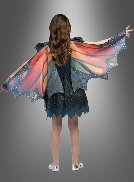 Ein Kind, das das farbenfrohe „Kleid mit Schmetterlingsflügel“ trägt – ein marineblaues Kleid mit bunten Schmetterlingsflügeln –, kombiniert mit silbernen Schuhen, steht mit ausgestreckten Armen vor einem grauen Hintergrund mit Farbverlauf. Dieses bezaubernde Kinderkostüm fängt die Magie der kindlichen Fantasie wunderbar ein.